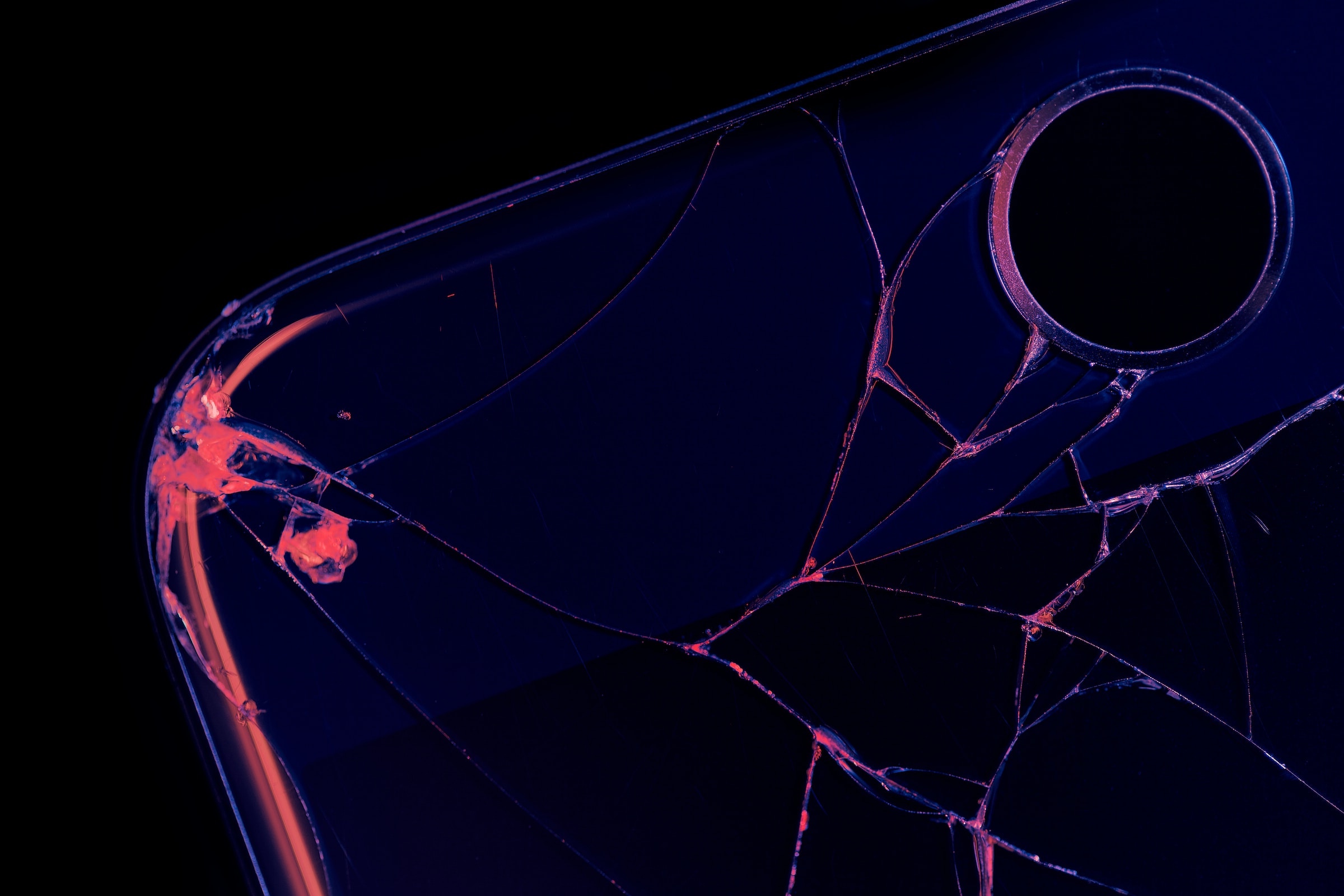A close-up image of a cracked phone showing the camera lense, in black and neon pink and purple A close-up image of a cracked phone showing the camera lense, in black and neon pink and purple